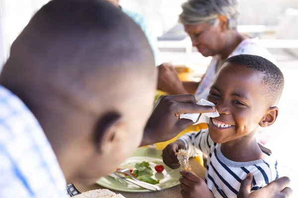 多人種の父は食卓で昼食をとりながらティッシュペーパーで笑顔の息子の鼻を掃除します 食べ物 変わらない 一緒に 子供時代と家庭の概念 — ストック写真