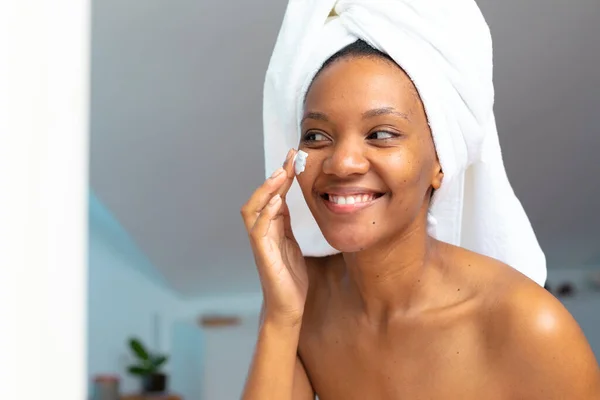 Χαμογελώντας Νεαρή Αφροαμερικανή Γυναίκα Κεφάλι Τυλιγμένο Πετσέτα Εφαρμογή Κρέμα Προσώπου — Φωτογραφία Αρχείου