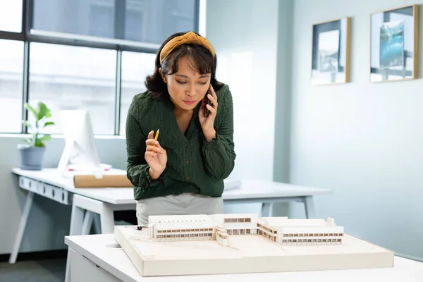Asian young architect talking on mobile phone while examining architectural model in office. Unaltered, workplace, creative business, design professional, development, architecture.
