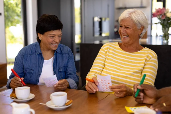 Vrolijke Biracial Senior Vriendinnen Met Koffie Koekjes Eettafel Spelen Bingo — Stockfoto