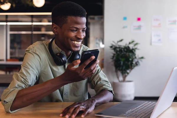 Smiling african american young businessman talking through smartphone speaker in office. Unaltered, creative business, wireless technology, voicemail, speech recognition.