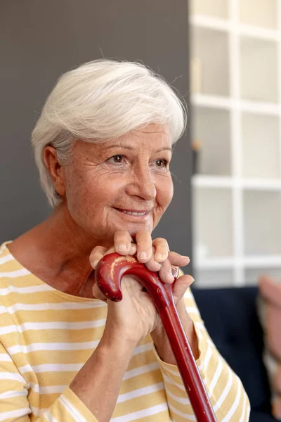 自宅のソファに腰掛けて笑顔で歩く棒を持つ白人のシニア女性 定年退職者のライフスタイルと生活コンセプト — ストック写真