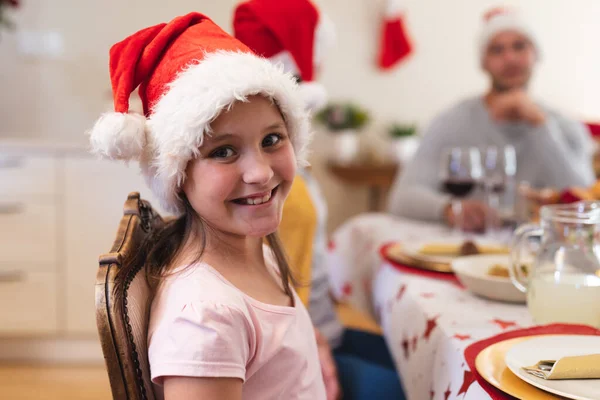 白人の少女の肖像画夕食のテーブルに座って サンタの帽子をかぶって カメラを見て笑顔で 背景に彼女の家族と一緒に 家族の時間クリスマスのお祝い — ストック写真