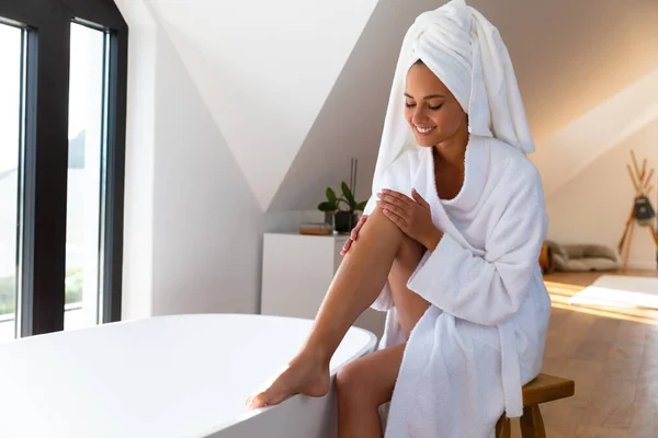 一名身穿浴衣的白人妇女坐在浴室浴缸边 在腿上涂了润肤霜 自我照顾 卫生和放松的概念 — 图库照片