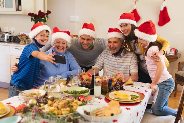 一緒に夕食のためにテーブルに座っている多世代の白人家族は サンタの帽子と顔のマスクを着て 自撮りと笑顔を取っています 家族の時間クリスマスのお祝い — ストック写真