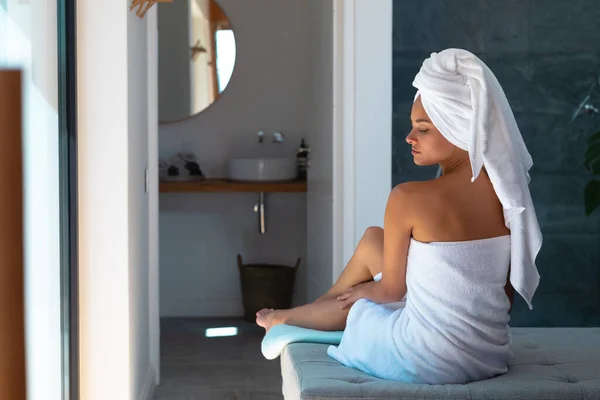 身穿浴衣的白人妇女坐在床上 在腿上涂了润肤霜 自我照顾 卫生和放松的概念 — 图库照片