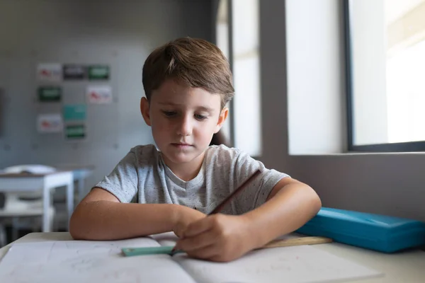 教室の机に座りながら本を書いている白人の小学生 変化のない教育子供時代学習学校の概念 — ストック写真