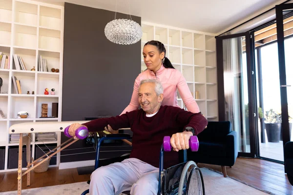 女性の出産理学療法士は 自宅でダンベルを持ち上げるには 白人のシニア男性を支援します 車椅子 変更されていない 理学療法 回復と障害 — ストック写真