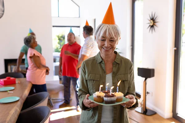 カップケーキやろうそくを持つアジア系のシニア女性や多人種の友人が背景で話しています 老人ホーム 誕生日 お祝い パーティー 一緒に 変更なし サポート 生活支援 — ストック写真