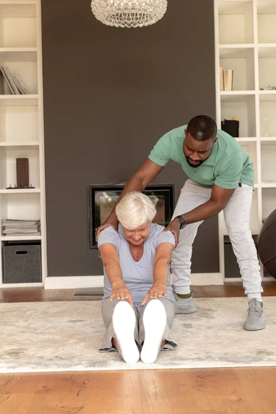 Afrikanisch Amerikanischer Physiotherapeut Hilft Kaukasischer Seniorin Sitzen Ihre Zehen Berühren — Stockfoto