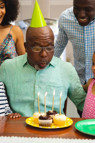 自宅で家族とお祝いしながら誕生日のろうそくを吹いて帽子をかぶって多人種のシニア男性 多世代 カップケーキ 変わらない 一緒に 甘い食べ物 楽しみ — ストック写真