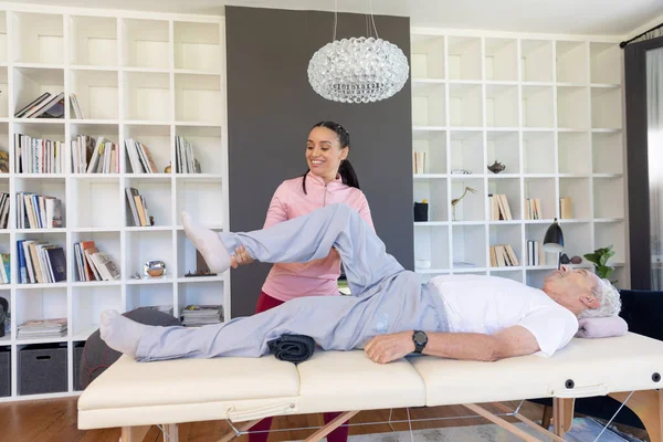 Ženský Biraciální Fyzioterapeut Natahující Nohu Staršího Bělocha Ležícího Doma Stole — Stock fotografie