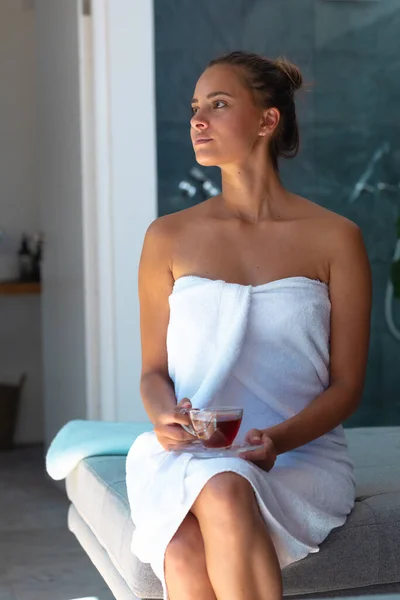 身穿浴衣的白种人年轻女子拿着茶杯坐在家里的床上 自我照顾 卫生和放松的概念 — 图库照片