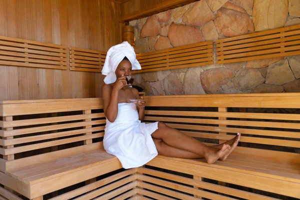 非洲裔美国妇女穿着浴衣 坐在桑拿的木制长椅上喝茶 未经改动的温泉 身体护理和幸福概念 — 图库照片
