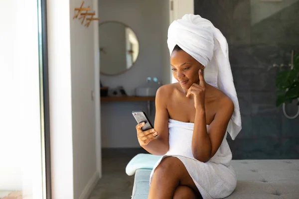自宅のベッドに座っているスマートフォンを使用してバスローブのアフリカ系アメリカ人女性 セルフケア リラクゼーションの概念 — ストック写真