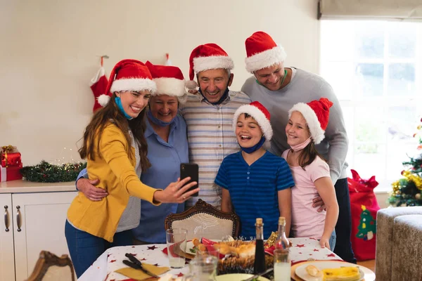 多代高加索人家庭一起站在餐桌前吃饭 戴着桑塔帽和面罩 带着自拍和微笑 优质家庭时间圣诞庆祝活动 — 图库照片