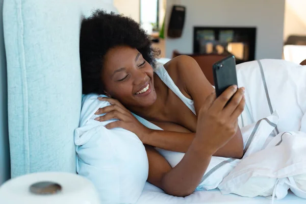 自宅のベッドに横たわっている間 スマートフォンを使用して若いアフリカ系アメリカ人のアフロ女性を笑顔 スペースをコピーします 変化のない ライフスタイル テクノロジーの概念 — ストック写真