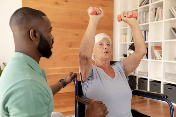 アフリカ系アメリカ人男性の理学療法士は ダンベルを使って運動をするために白人の高齢女性を助ける 理学療法とリハビリテーションの概念 — ストック写真