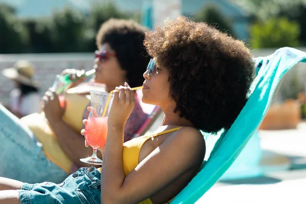 在泳池派对上 戴着太阳镜的年轻女孩躺在甲板椅子上喝鸡尾酒 非洲头发 社交活动 享乐和周末活动 — 图库照片