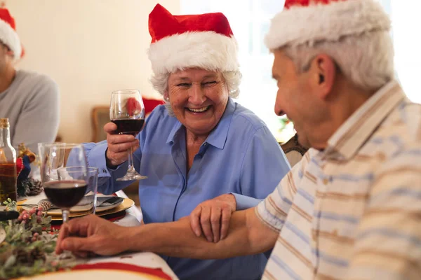 资深的高加索夫妇一起坐在餐桌前吃饭 戴着桑塔帽 相互望着对方 微笑着 女人举着酒杯 优质家庭时间圣诞庆祝活动 — 图库照片