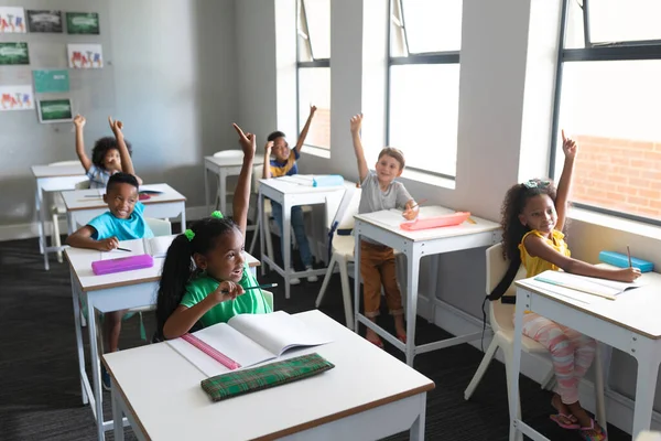 Uśmiechnięci Wielorasowi Uczniowie Szkoły Podstawowej Podniesionymi Rękami Siedzący Biurkiem Klasie — Zdjęcie stockowe