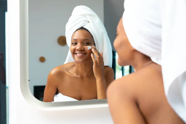 Sorrindo Jovem Afro Americana Aplicando Creme Facial Enquanto Olha Espelho — Fotografia de Stock