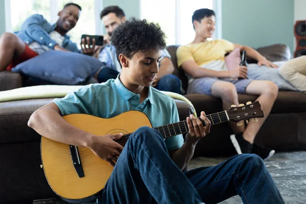 男性の友人は自宅でソファの上でリラックスしながら 多人種の若い男はギターを演奏します 変更されていない 社会的な集まり 楽しみと週末の活動 — ストック写真