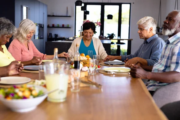 Multiracial senior friends with food on dining table holding hands and saying grace in nursing home. Praying, togetherness, lunch, unaltered, support, assisted living and retirement concept.