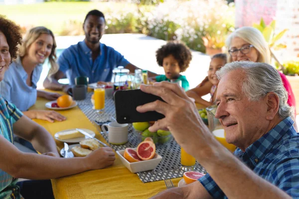 Улыбающийся Пожилой Человек Делает Селфи Смартфоном Мультирасовой Семьей Завтраком Дом — стоковое фото