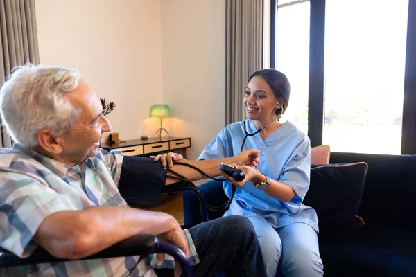 Smiling multiracial female doctor examining senior man's blood pressure with stethoscope and gauge. Home, sick, unaltered, hypertensive, healthcare, patient, retirement, recovery, disability.