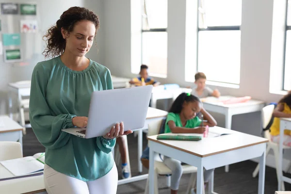 白人の若い女性教師は 多人種の小学生を背景にラップトップを使用しています 変化のない教育子供時代学習無線技術職業学校の概念 — ストック写真