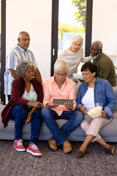 在养老院里 与爆米花一起观看数字平板电脑比赛的多种族资深朋友 无线技术 辅助生活和退休 — 图库照片