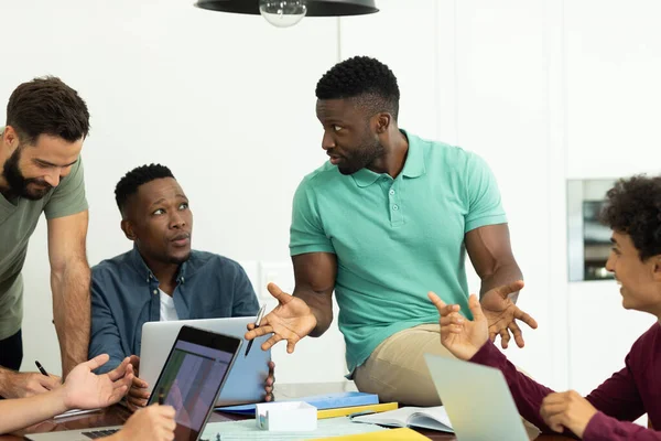 Businessman gesturing and explaining strategies to multiracial male coworkers while sitting on desk. Office, planning, wireless technology, unaltered, creative business and teamwork concept.