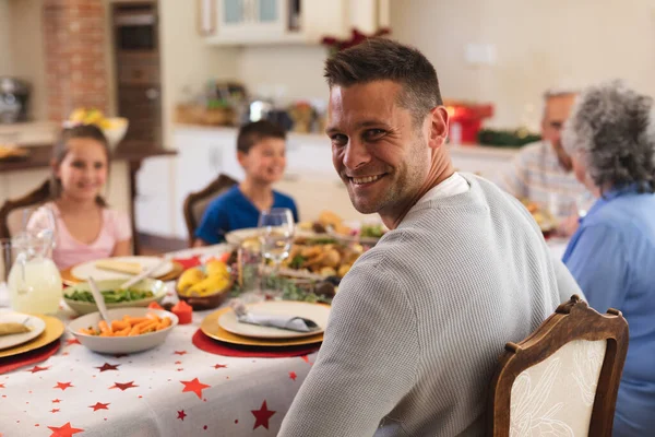 一个高加索人的画像 他坐在餐桌旁吃饭 看着相机 面带微笑 背后是他的家人 优质家庭时间圣诞庆祝活动 — 图库照片