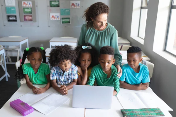 アフリカ系アメリカ人の小学生がノートパソコンを使って机の上に立つ白人の若い女性教師 変化のない教育子供時代教育学習無線技術学校の概念 — ストック写真