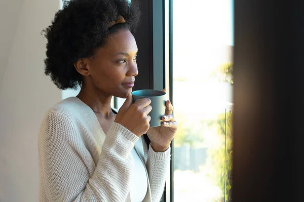 アフリカ系アメリカ人の若い女性が家で窓から外を眺めながらコーヒーカップを持っている リラクゼーションのコンセプト — ストック写真