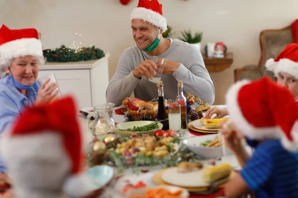 一代又一代的高加索人家庭坐在桌旁 面带微笑 戴着桑塔帽和面罩 优质家庭时间圣诞庆祝活动 — 图库照片