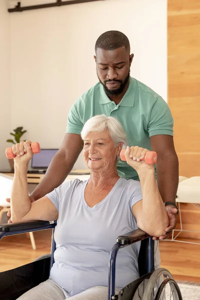 アフリカ系アメリカ人男性の理学療法士は ダンベルを使って運動をするために白人の高齢女性を助ける 理学療法とリハビリテーションの概念 — ストック写真
