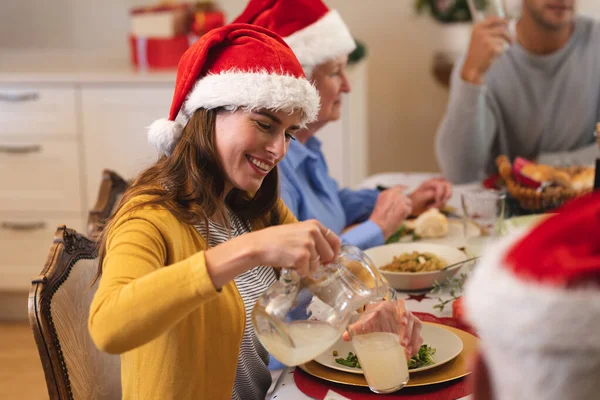 白人女性は夕食のためのテーブルに座って サンタの帽子をかぶって 背景に彼女の家族と一緒にガラスと笑顔にレモネードを注ぐ 家族の時間クリスマスのお祝い — ストック写真