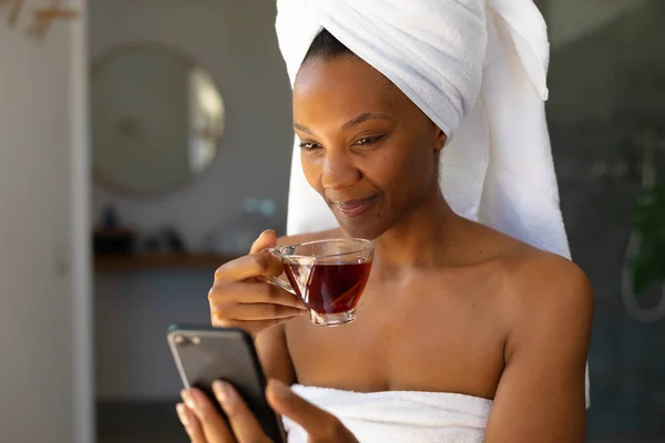 穿着浴衣的非洲裔美国妇女在家里用智能手机拿着茶杯 自我照顾 卫生和放松的概念 — 图库照片