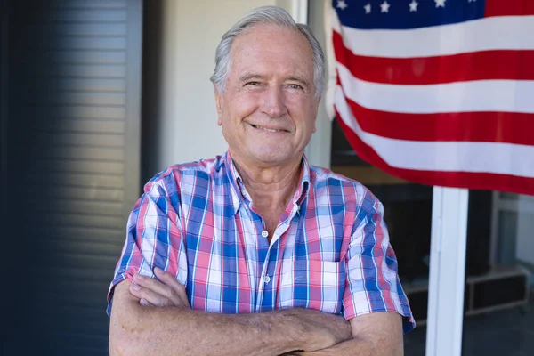 Portrait of caucasian senior man with arms crossed standing with flag of america in background. Copy space, house, smiling, unaltered, lifestyle, retirement and patriotism concept.