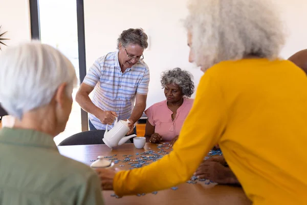白种人老人在养老院为多种族朋友提供咖啡 玩拼图游戏 头脑风暴 辅助生活和退休 — 图库照片