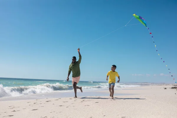 晴れた日にビーチでカイトを飛んでいる間に実行している遊び心のあるアフリカ系アメリカ人の父と息子は スペースをコピーします 変更されていない ライフスタイル 楽しさと休日の概念 — ストック写真
