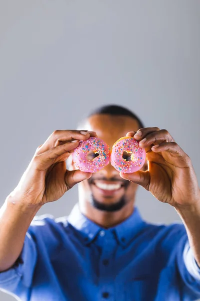 stock image African american man holding pink donuts in front of face with copy space against gray background. unaltered, unhealthy eating and sweet food concept.