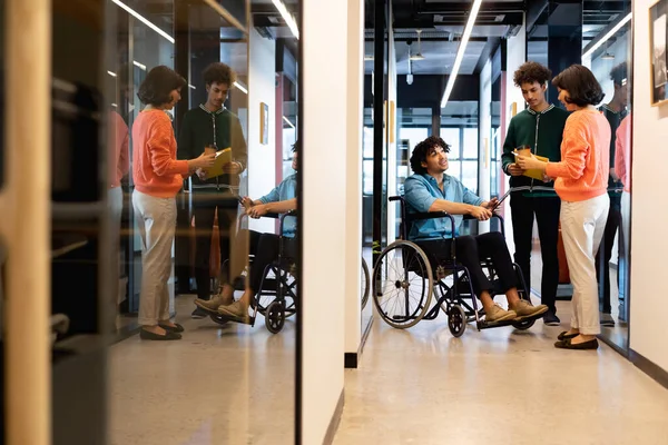 Multiracial colleagues discussing with young businessman sitting in wheelchair at office corridor. Unaltered, teamwork, creative business, workplace, diversity, disability, support.