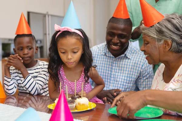 家で家族と誕生日を祝いながら 帽子を吹いてろうそくを身に着けている多人種の興奮した女の子 パーティー 変更されていない 一緒に 子供時代 カップケーキ 楽しみと退職 — ストック写真