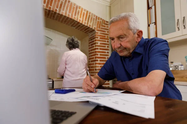 年长的高加索人男人呆在家里 坐在厨房里 使用笔记本电脑 看文件 而他的妻子则站在后面 检疫封闭过程中的分离分离物 — 图库照片