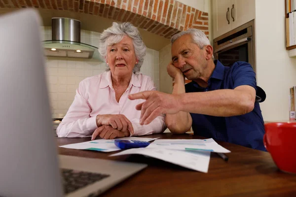 高齢者の白人カップル一緒に家で時間を過ごします キッチンに座って ノートパソコンを使用して 書類を見て — ストック写真