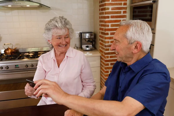 高齢者の白人のカップルは 一緒に家で時間を過ごし キッチンに座ってスマートフォンと笑顔を使っています コロナウイルスCovid 19隔離ロックダウン中に隔離 — ストック写真