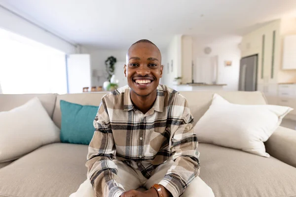 Stock image Portrait of smiling african american young man sitting on sofa and looking at camera in living room. Copy space, unaltered, happy, relaxation, lifestyle and home concept.